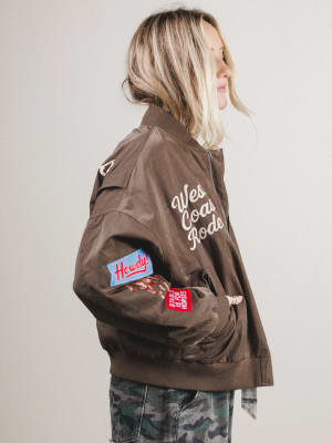 Woman wearing a brown jacket with patches and text, standing against a plain background