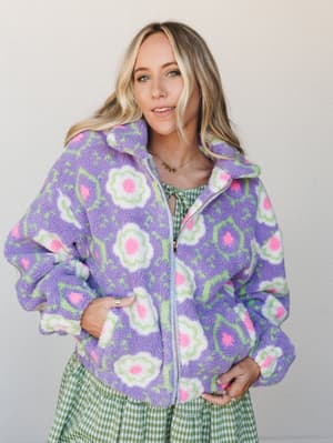 A sherpa zip-up jacket in cheerful purple with oversized retro floral print, layered over a green gingham dress — the kind of outfit that looks like it was styled by someone who genuinely loves getting dressed.