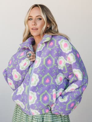 A bold, oversized floral sherpa zip-up jacket in lavender purple with pink and white blooms — styled with green gingham bottoms and stacked turquoise rings for a retro-playful, cozy-cool look shot against a clean light backdrop.