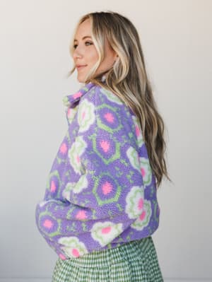 A model shot from behind-ish, glancing back over her shoulder in a chunky floral sherpa jacket in purple, hot pink, and sage green — layered over a green gingham skirt, the whole look giving very 'color theory homework, but make it cozy.'