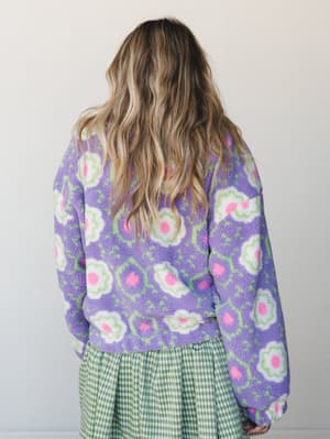 Shot from behind, a model with loose wavy hair wears a cozy sherpa pullover in bold purple with an oversized retro floral print, paired with a green gingham skirt — the kind of pattern-mixing that feels intentional and a little joyful.