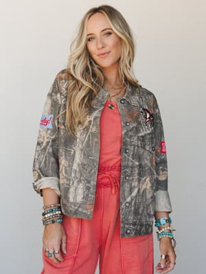 A camo utility jacket covered in playful patches — horse, 'Howdy,' and more — layered over a coral matching set, styled with stacked turquoise and beaded bracelets for an effortlessly Western-boho look.