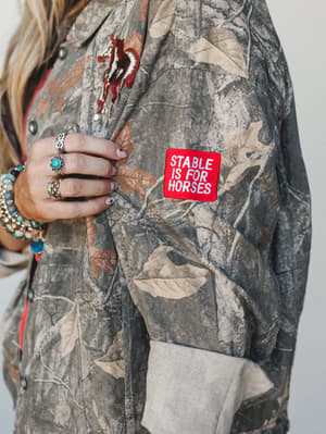 A close-up detail shot of a camo jacket sleeve with a bold red 'Stable Is For Horses' embroidered patch and a horse graphic embroidery on the chest, styled with stacked turquoise and silver rings and boho bead bracelets — Western irreverence at its b