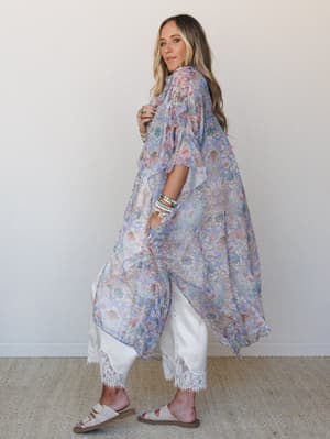 A flowy, floor-length floral kimono in soft periwinkle and blush tones layered over white wide-leg pants with lace trim — the kind of outfit that feels effortless but completely intentional.