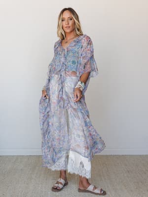 A flowy periwinkle floral kimono worn open over white wide-leg lace-trim pants, styled with stacked bracelets and sandals against a soft neutral studio wall — effortlessly dreamy and put-together at the same time.