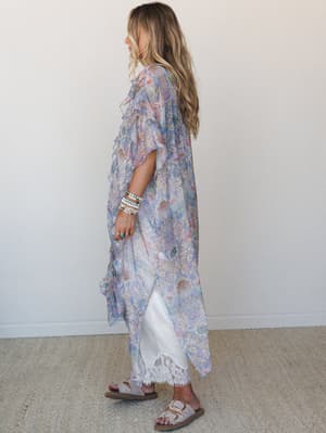 A flowy, floor-length paisley kimono in soft periwinkle and blush layers over a white slip dress with lace hem, styled with beaded bracelets and birkenstock sandals — the kind of outfit that looks effortless but definitely wasn't an accident.