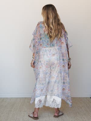 Shot from behind, a model layers a sheer periwinkle paisley kimono over white lace-trim pants, the whole look feeling like something you'd wear to wander a coastal market with bracelets stacked to your elbow.