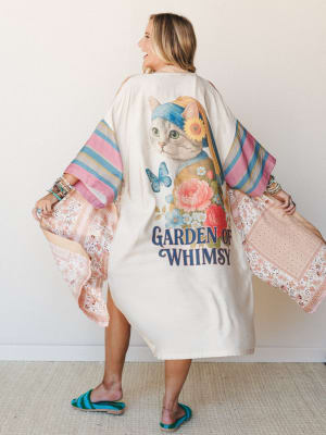 A model shows off the back of a cream kimono featuring a whimsical cat graphic surrounded by painted florals, butterflies, and 'Garden of Whimsy' lettering, with wide sleeves in mixed pink stripe and floral patchwork panels — playful, personal, and c