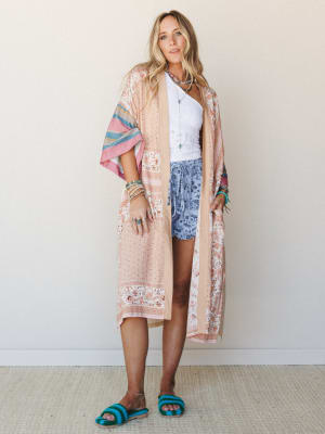 A long floral kimono in peachy natural tones with striped sleeves is layered over a white lace crop top and periwinkle floral shorts, styled with turquoise beaded sandals and stacked boho jewelry — the whole look feels like a summer afternoon that re