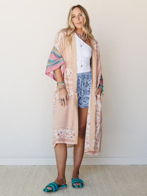 A long floral kimono in peachy natural tones with striped sleeves is layered over a white lace crop top and periwinkle floral shorts, styled with turquoise beaded sandals and stacked boho jewelry — the whole look feels like a summer afternoon that re