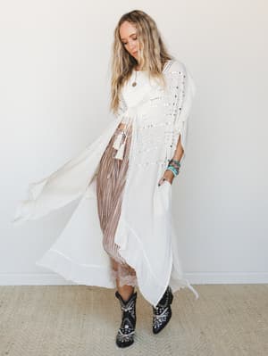 A flowy vanilla cream kimono with mirror stud embellishment moves mid-stride against a clean white wall, styled over a cropped white top, pleated mauve midi skirt, and embroidered black western boots — the kind of outfit that feels effortless and int