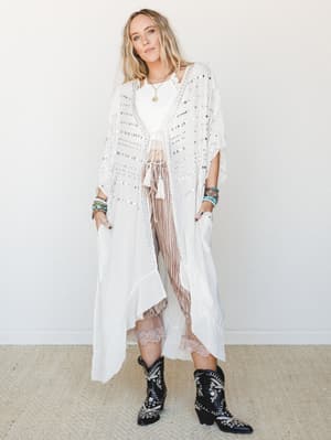 A long white studded kimono layered over a white crop top and striped wide-leg pants with a lace hem, styled with embroidered black cowboy boots and stacked turquoise bracelets — free-spirited and effortlessly put-together.