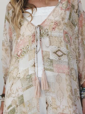 A close-up torso shot of a sheer patchwork-print kimono layered over a white top, with delicate tassel tie closure at the neckline — soft, romantic, and full of that lived-in boho energy.