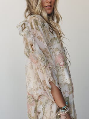 A flowy patchwork-print kimono in soft blush and olive tones photographed from the side, styled with a stack of beaded bracelets — the kind of piece that looks equally at home over a swimsuit or a sundress.