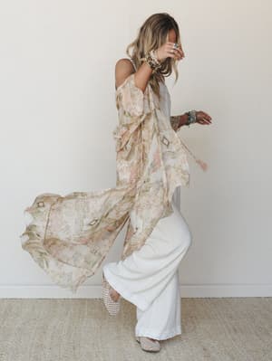 A flowy printed kimono in soft blush and sage tones catches mid-movement over wide-leg white pants, stacked boho jewelry finishing the look — the whole thing feels like a good afternoon with nowhere to be.