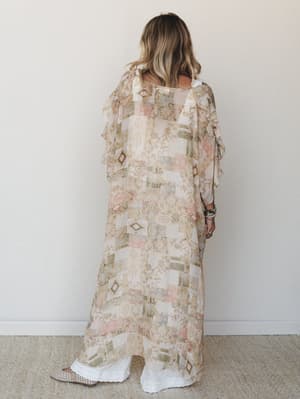 A model shot from behind wearing a floor-length sheer kimono in a warm patchwork-style mixed print — layered over wide-leg white pants with stacked turquoise bracelets adding just the right amount of personality.