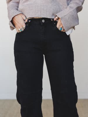 A close-up torso shot of black barrel-leg jeans styled with a soft lavender-gray ribbed sweater, hands tucked into pockets and stacked with silver and turquoise rings — the kind of effortless pairing that looks like it took zero effort and exactly th