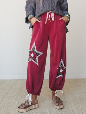 A relaxed wide-leg jogger in deep magenta with layered star patchwork appliqués on each leg, styled with a chunky cable-knit sweater, stacked beaded bracelets, and leopard print platform sandals — the kind of outfit that's effortlessly expressive wit