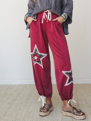 A relaxed pair of magenta jogger-style pants with lace-trimmed star patchwork appliqués on each leg, styled with a chunky grey cable knit sweater, stacked boho bracelets, and espadrille platform sandals — the kind of outfit that looks effortless but