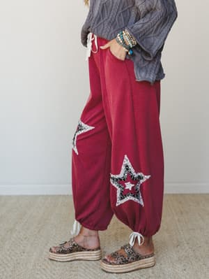A cropped side-profile shot of relaxed wide-leg pants in deep magenta, styled with a chunky cable-knit sweater and platform sandals — the star patchwork appliqués and lace-trim details give these a playful, worn-in boho energy.