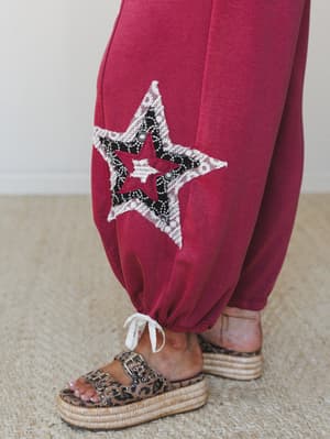 A close-up detail shot of magenta balloon-hem pants featuring a layered star appliqué in lace and black fabric, styled with leopard print platform sandals on a woven rug.