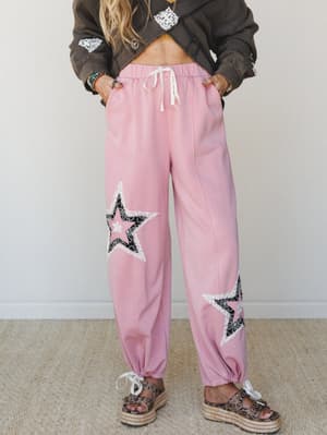 Pink wide-leg jogger pants with layered star appliqués in black lace and cream trim, styled with a cropped charcoal jacket and platform sandals — playful and boho with a retro edge.