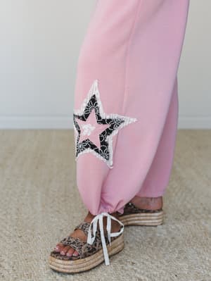 A close-up detail shot of wide-leg pink pants with a statement star appliqué in black lace and raw fringe edges, paired with leopard print platform espadrille sandals — the kind of detail that makes someone stop mid-scroll.