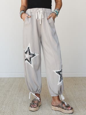 Relaxed taupe jogger-style pants with lace and embroidered star appliqués on each leg, tied off at the ankles, styled with stacked bracelets and espadrille platform sandals for that effortlessly expressive everyday look.