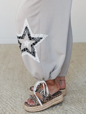 A close-up detail shot of the hem of a taupe wide-leg pant, featuring a layered star appliqué in black lace and cream lace trim, paired with leopard print platform espadrille sandals tied with a lace ribbon.