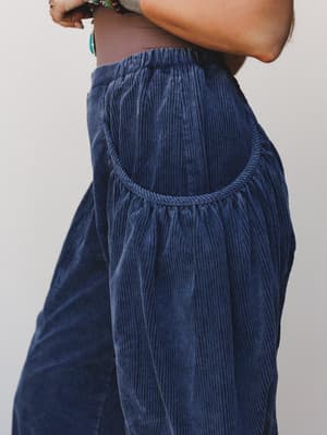 A close-up detail shot of deep navy corduroy pants with a gathered, rounded side pocket and elastic waistband — the texture is rich and tactile, styled with layered boho bracelets peeking into frame.