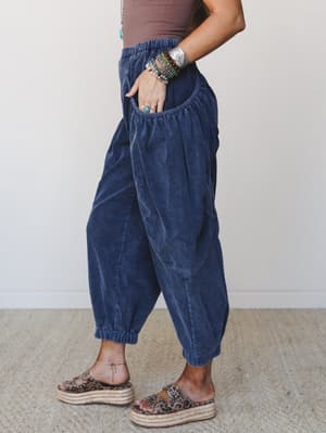 A side-profile shot of wide-leg corduroy barrel pants in denim blue, styled with a brown tank, leopard print platform sandals, and a stack of boho bracelets — the kind of outfit that looks effortless but is very much intentional.