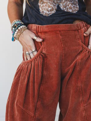 A close-up detail shot of rust-orange wide corduroy barrel pants styled with a washed graphic tee, hands tucked in gathered pockets and layered with beaded bracelets and silver rings — the kind of outfit that feels pulled-together without trying.