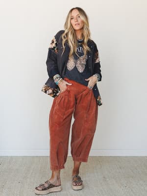 A full-body lifestyle shot of a model in rust corduroy barrel pants layered with a graphic tee and a floral-patched black denim jacket — the kind of outfit that looks like it came together effortlessly but definitely did not.