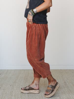 A side-profile shot of rust corduroy barrel-leg pants styled with a washed black graphic tee, leopard print platform sandals, and a stacked mix of turquoise and silver bracelets — earthy, relaxed, and full of personality.