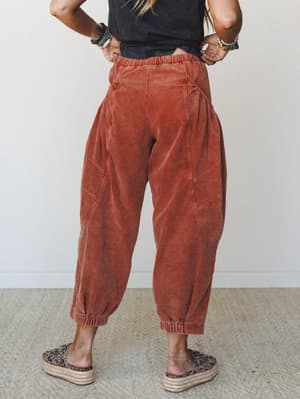 A back-view shot of barrel-fit corduroy pants in a warm rustic orange, styled with a cropped black tee, layered bead bracelets, and leopard print platform espadrilles — relaxed and expressive without trying too hard.