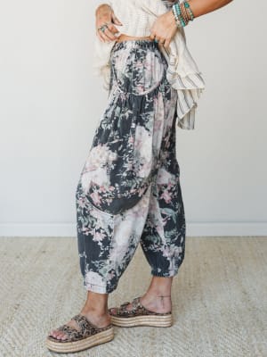 A side-angle shot of floral barrel-leg pants in faded charcoal with blush and sage blooms, styled with a cream ruffled top, stacked turquoise bracelets, and leopard print platform sandals — effortlessly boho with just enough edge.