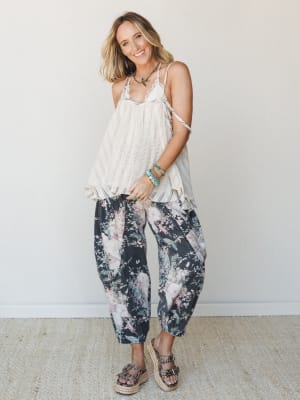 A smiling model pairs a breezy cream lace-trim cami with dark floral barrel-leg pants and studded platform espadrilles — the kind of outfit that looks effortless but clearly wasn't an accident.