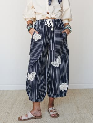 Wide-leg barrel pants in navy pinstripe steal the whole show — lace butterfly and floral appliqués scattered across patch pockets, paired with a knotted cream top and stacked bracelets for that effortlessly collected, boho-meets-nautical feel.