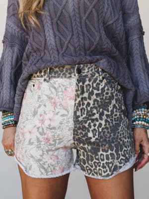 A close-up shot of mixed-print denim cutoff shorts — half floral, half leopard — paired with a chunky cable-knit sweater and stacked beaded bracelets, giving off that perfectly imperfect, collected-over-time energy.