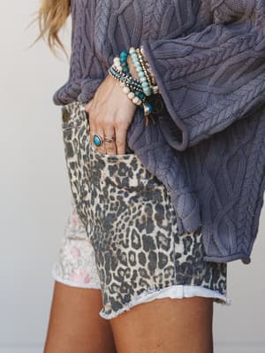 A close-up of a mixed-print short — leopard front, floral panel — styled with an oversized cable knit sweater in dusty blue and a stack of boho bracelets and stone rings that do all the talking.