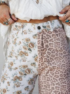 A close-up of mixed-print wide leg jeans styled with a cropped white lace and gauze top — one leg in a vintage floral, the other in a soft leopard print, both in sun-bleached neutrals that feel effortlessly collected rather than matchy.