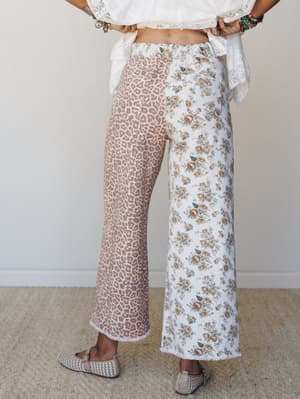 A back-facing model styled in mixed-print wide-leg jeans — one leg in blush leopard, one in cream vintage floral — paired with a white lace crop top and woven flats, shot against a clean studio wall with an effortlessly boho energy.