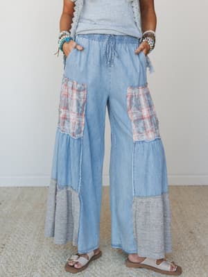 Wide-leg patchwork pants in light chambray denim with faded plaid contrast panels, raw-edge tiered seams, and a cozy knit hem — styled with stacked beaded bracelets and strappy sandals for a free-spirited, effortlessly collected look.