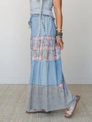 A tiered maxi skirt (or wide-leg pant) in light denim and mixed fabrics — chambray, faded plaid, and heathered grey — stacked in panels with lace trim at the waist and a rhinestone star appliqué, styled with stacked beaded bracelets and white slide s