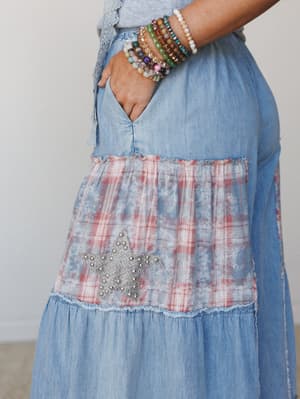 A close-up detail shot of light denim tiered pants featuring a bleached plaid patchwork panel with a studded star appliqué, worn with a stack of beaded bracelets — the kind of piece that feels like it has a story already.