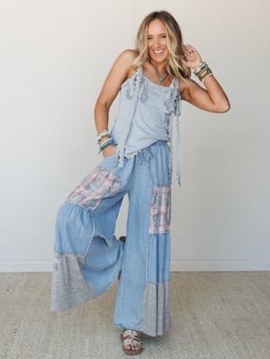 A smiling model in a breezy studio setting wears ultra-wide-leg patchwork denim pants with a lace-trimmed cami top, layered bracelets, and sandals — the kind of outfit that looks effortless but is definitely doing a lot.