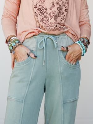 A close-up mid-shot of wide-leg sweatpants in a washed sage green paired with a blush long-sleeve graphic tee, hands tucked in pockets and layered beaded bracelets adding that effortless personal-style energy.