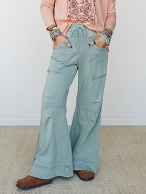 Wide-leg flare sweatpants in a washed sage tone styled with a peachy graphic tee, stacked turquoise bracelets, and brown suede boots — the kind of outfit that looks effortless but was definitely thought about.