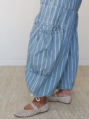 A close-up detail shot of wide-leg striped pants with a generous patch pocket and elasticized waist panel, paired with woven Mary Jane flats on a jute rug — effortless and a little unexpected.