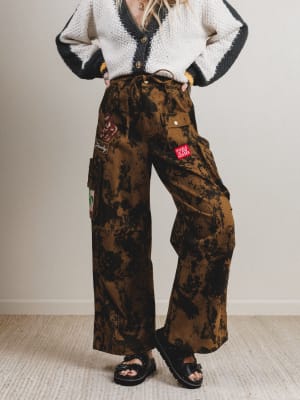 Person wearing a patterned cardigan and camouflage pants with patches on a plain background